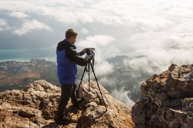 Bulutların, şehrin ve denizin üstündeki yüksek bir dağın tepesinde tripod ve kamerayla duran bir adam. Profesyonel fotoğrafçı kayalık zirvede DSR ayarlarını ayarlıyor.