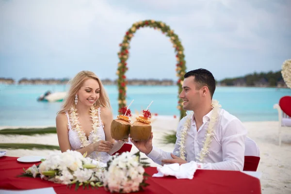 O noivo e a noiva bebem coquetéis de coco na mesa de casamento com lei de flores na praia da ilha tropical nas Maldivas. Oceano azul-turquesa e arco cerimonial no fundo. Conceito de lua de mel . — Fotografia de Stock