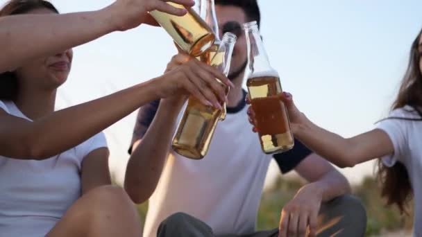 Group of friends have fun toasting, drinking beer, relaxing on sea beach at sunset in slow motion. Young men, women enjoy beverage sitting on a sand on warm summer evening party. People with lemonade. — Stock Video