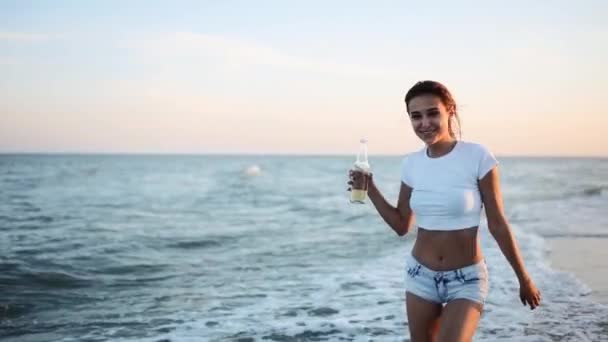 Mulher bonita andando ao longo do litoral na praia de areia se divertindo com garrafa de cerveja. Menina equipada atraente levanta as mãos, goza de fredom e férias de verão perto da costa do oceano, bebendo bebida. Movimento lento. — Vídeo de Stock