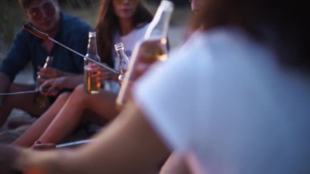 Friends sit around bonfire cheering and toasting beer bottles, playing guitar, and frying sausages on sandy beach. Young group of men and women drink beverages, singalong. Guy playing guitar in dusk. — Stock Video