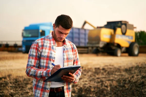 Farmer controls loading wheat from harvester to grain truck. Driver holding clipboard, keeping notes, cargo counting. Forwarder fills in consignment waybills. Agricultural commodities logistics.