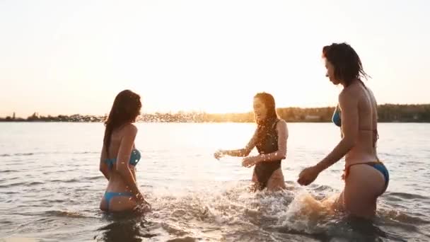 Le ragazze bagnate felici in bikini corrono in mare e giocano spruzzando acqua l'un l'altro al tramonto. Le amiche allegre si divertono a fare schizzi nello stagno. Giovani donne andare a nuotare, fare il bagno nel lago. Rallentatore. — Video Stock