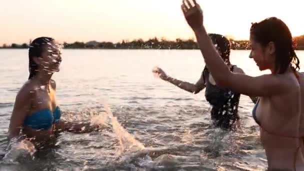 Le ragazze bagnate felici in bikini corrono in mare e giocano spruzzando acqua l'un l'altro al tramonto. Le amiche allegre si divertono a fare schizzi nello stagno. Giovani donne andare a nuotare, fare il bagno nel lago. Rallentatore. — Video Stock