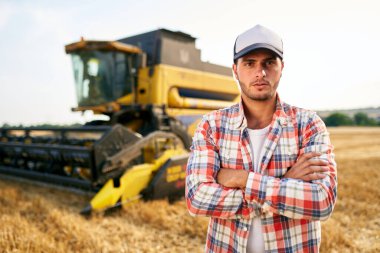 Göğsünde elleri çapraz olan gururlu hasatçı makinist portresi. Çiftçi kombinasyonunda duruyor. Agronomist kameraya bakıyor. Hasat edilen buğday tarlaları üzerinde çalışan çiftçi..