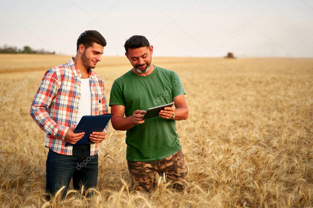 Two farmers stand in wheat stubble field, discuss harvest, crops. Senior agronomist with touch tablet pc teaches young coworker. Innovative tech. Precision farming with online data management soft.