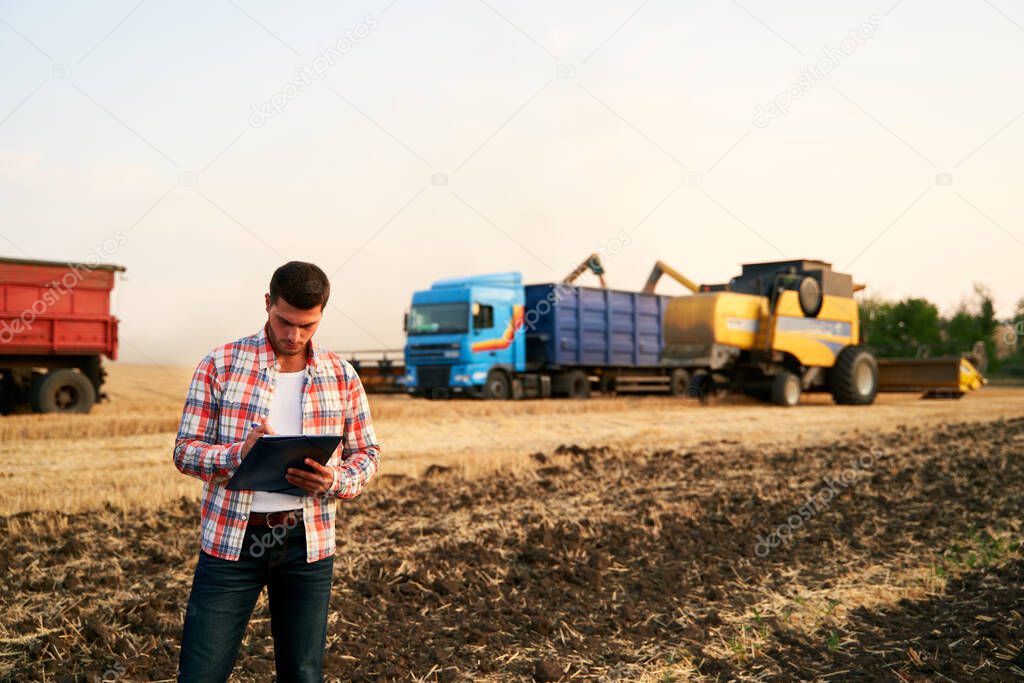 Farmer controls loading wheat from harvester to grain truck. Driver holding clipboard, keeping notes, cargo counting. Forwarder fills in consignment waybills. Agricultural commodities logistics.