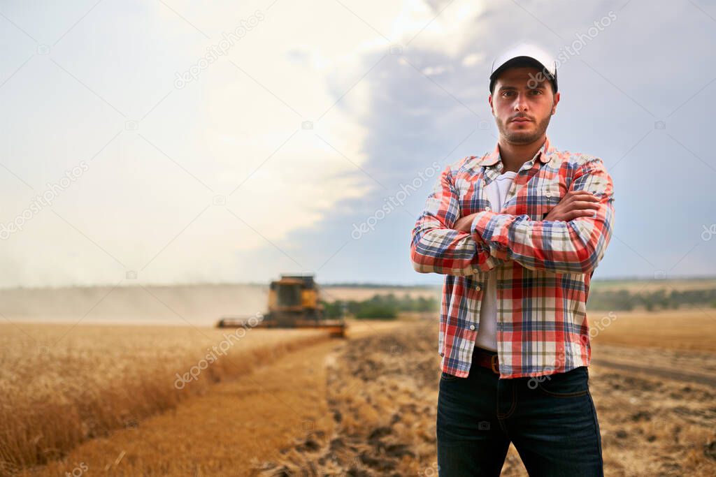 Happy farmer proudly standing in a field with arms crossed on chest. Combine harvester crop rich wheat harvest on background. Agronomist wearing flannel shirt, looking at camera on a farmland.