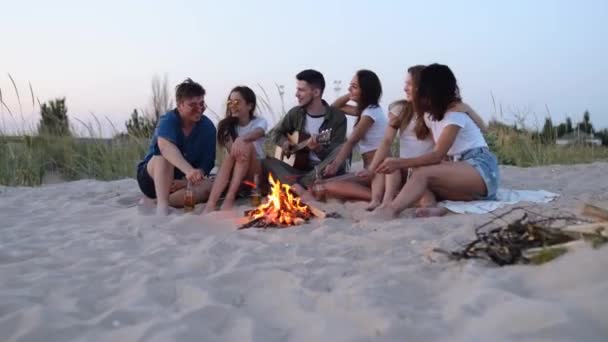 Friends sit around bonfire, drink beer, sing to guitar, fry sausages on sandy beach. Young mixed race group of men and women with beverage singalong, bearded guy playing guitar on party in the dusk. — Stock Video