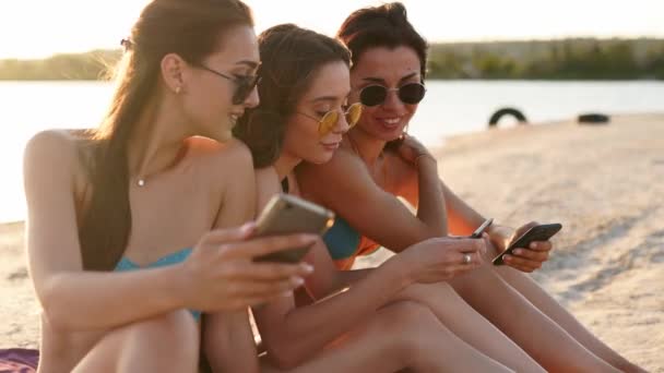 Groupe de filles millénaires utilisant des smartphones assis ensemble sur une serviette de plage près de la mer au coucher du soleil d'été. Jeunes femmes dépendantes des téléphones portables intelligents. Toujours connecté génération communiquer via Internet. — Video