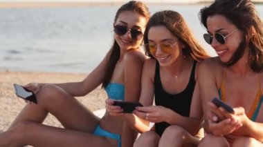 Bin yıllık kızlar yaz günbatımında deniz kenarında plaj havlusunda birlikte oturuyorlar. Cep telefonu bağımlısı genç kadınlar. Her zaman bağlı nesil internet aracılığıyla iletişim.