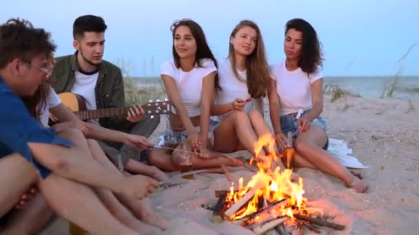 Gli amici si siedono intorno al falò, bevono birra, cantano alla chitarra, friggono salsicce sulla spiaggia sabbiosa. Giovane razza mista di uomini e donne con bevanda singalong, ragazzo barbuto suonare la chitarra alla festa al crepuscolo. — Video Stock