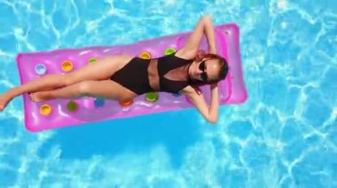 Yüzme havuzunda bikinili güzel bir kız şişme pembe yatakta dinleniyor. Mayo giymiş, bronzlaşmış bir kadın. Lüks oteldeki mavi suda yüzen bir kadın. Gökyüzü, yukarıdan görünüm.