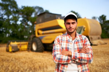 Göğsünde elleri çapraz olan gururlu hasatçı makinist portresi. Çiftçi kombinasyonunda duruyor. Agronomist kameraya bakıyor. Hasat edilen buğday tarlaları üzerinde çalışan çiftçi..