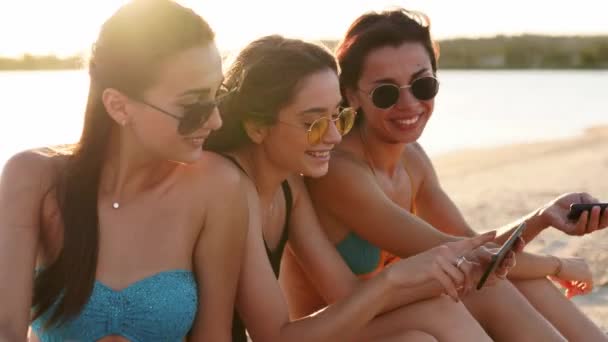 Grupo de meninas milenares usando smartphones sentados juntos na toalha de praia perto do mar no pôr do sol de verão. Jovens viciadas em telemóveis inteligentes. Geração sempre conectada comunicar via internet . — Vídeo de Stock