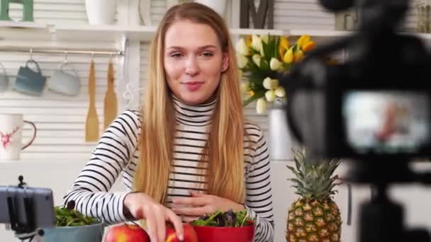 Camera scherm opname - focus transitie - Food blogger koken verse veganistische salade van fruit in de keuken studio, filmen tutorial. Vrouwelijke influencer houdt appel, ananas, praat over gezond eten. — Stockvideo