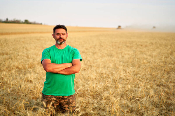 Happy farmer proudly standing in wheat field with arms crossed on chest. Agronomist wearing corporate uniform, looking at camera on farmland. Rich harvest of cultivated cereal crop. Harvesting season.