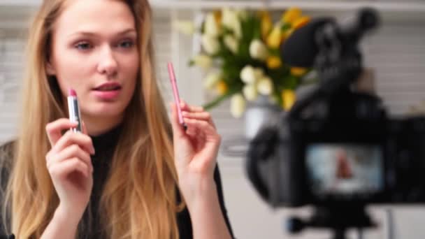 Vlogger θηλυκό εφαρμόζει κραγιόν στα χείλη. Beauty blogger γυναίκα μαγνητοσκόπηση καθημερινή make-up ρουτίνας φροντιστήριο στην κάμερα στο τρίποδο. Influencer ξανθιά κοπέλα ζωντανή μετάδοση καλλυντικών σύγκριση προϊόντων στο στούντιο — Αρχείο Βίντεο