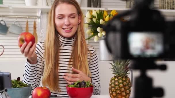 Grabación de pantalla de cámara - transición de enfoque - Blogger de alimentos cocinando ensalada vegana fresca de frutas en el estudio de cocina, filmando tutorial. Influenciadora femenina sostiene manzana, piña, habla de una alimentación saludable . — Vídeos de Stock