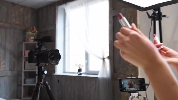 Vlogger hembra aplica lápiz labial en los labios. Belleza blogger mujer filmación diaria de maquillaje tutorial de rutina en la cámara en trípode. Influencer chica rubia streaming en vivo cosméticos revisión del producto en el estudio en casa — Vídeo de stock