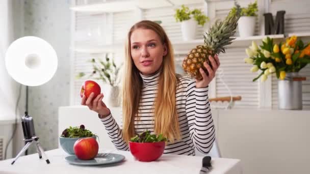 Yemek blogcusu mutfak stüdyosunda taze meyve salatası pişiriyor video kanalı için özel ders kameraları önünde çekim yapıyor. Kadın nüfuzlu kişi elmayı, ananası tutuyor ve sağlıklı beslenmekten bahsediyor. Fructorianism — Stok video