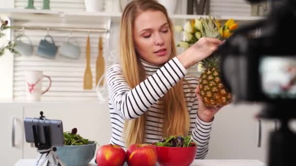 Grabación de pantalla de cámara - transición de enfoque - Blogger de alimentos cocinando ensalada vegana fresca de frutas en el estudio de cocina, filmando tutorial. Influenciadora femenina sostiene manzana, piña, habla de una alimentación saludable . — Vídeo de stock