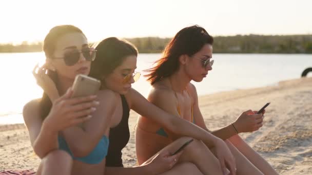 Grupo de chicas milenarias usando teléfonos inteligentes sentados juntos en la toalla de playa cerca del mar en el atardecer de verano. Mujeres jóvenes adictas a los teléfonos inteligentes móviles. Generación siempre conectada comunicarse a través de Internet. — Vídeo de stock