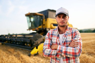 Göğsünde elleri çapraz olan gururlu hasatçı makinist portresi. Çiftçi kombinasyonunda duruyor. Agronomist kameraya bakıyor. Hasat edilen buğday tarlaları üzerinde çalışan çiftçi..