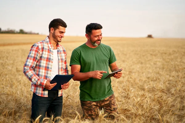 Two farmers stand in wheat stubble field, discuss harvest, crops. Senior agronomist with touch tablet pc teaches young coworker. Innovative tech. Precision farming with online data management soft.