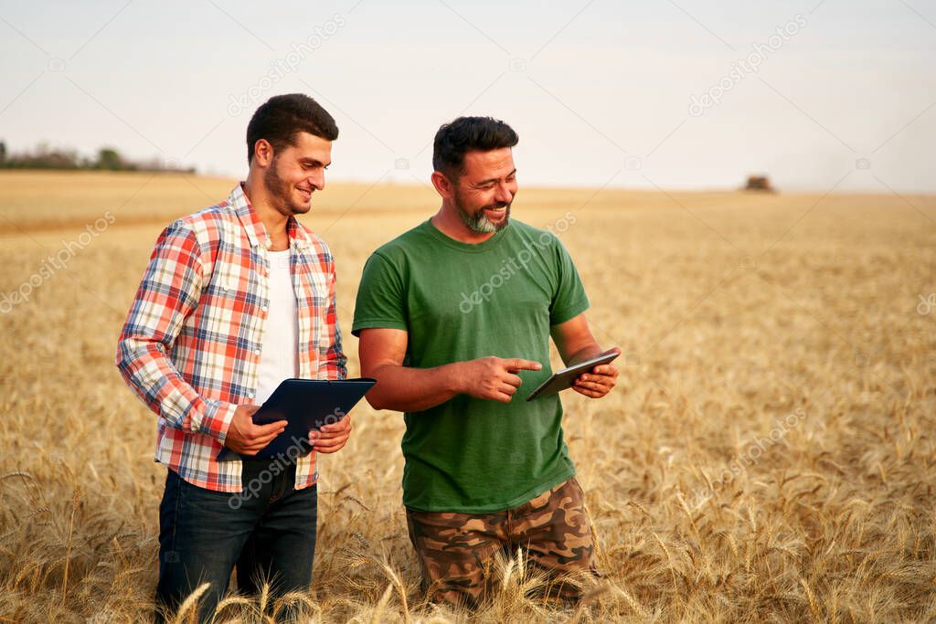 Two farmers stand in wheat stubble field, discuss harvest, crops. Senior agronomist with touch tablet pc teaches young coworker. Innovative tech. Precision farming with online data management soft.