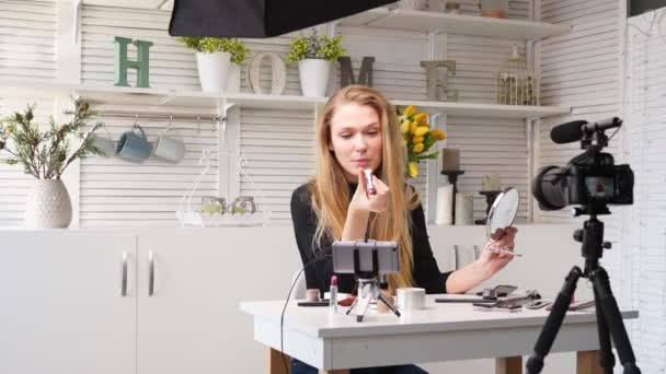 Vlogger fêmea aplica batom nos lábios. Mulher blogueira de beleza filmando tutorial de rotina de maquiagem diária na câmera no tripé. Influenciador menina loira ao vivo streaming cosméticos revisão do produto em estúdio em casa — Vídeo de Stock