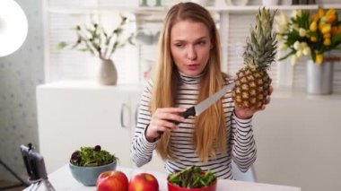 Yemek blogcusu mutfak stüdyosunda taze meyve salatası pişiriyor video kanalı için özel ders kameraları önünde çekim yapıyor. Kadın nüfuzlu kişi elmayı, ananası tutuyor ve sağlıklı beslenmekten bahsediyor. Fructorianism