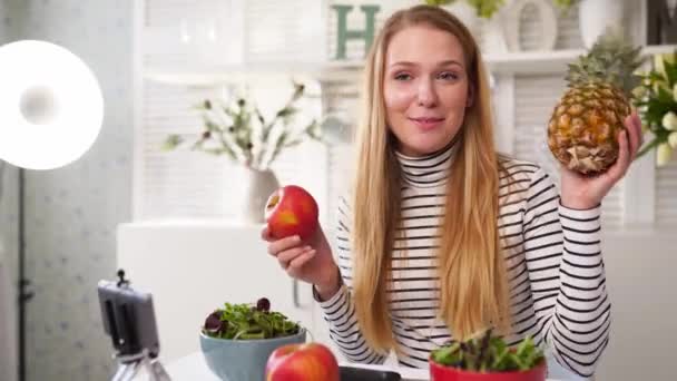 Yemek blogcusu mutfak stüdyosunda taze meyve salatası pişiriyor video kanalı için özel ders kameraları önünde çekim yapıyor. Kadın nüfuzlu kişi elmayı, ananası tutuyor ve sağlıklı beslenmekten bahsediyor. Fructorianism — Stok video
