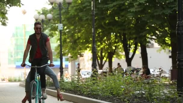 Een jong stel dat samen op de fiets rijdt in het park. Romantisch vriendje fietsen met gelukkig vriendin op cargo rack in de stad straat. Vrouw knuffels man hebben plezier rijden op de fiets in tandem. Langzame beweging. — Stockvideo