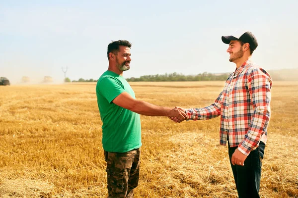 Agricultor e agrônomo apertando as mãos no campo de trigo após acordo. Conceito de contrato de negócio agrícola. Agricultor corporativo e fazendeiro proprietário negociar com aperto de mão. Colheitadeiras colheitadeiras colheita . — Fotografia de Stock