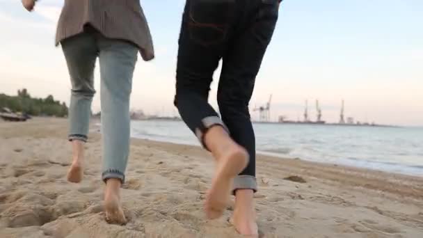 Hátsó alacsony szög kilátás romantikus pár láb futó homok fogja a kezét a tengerparton, közel a tenger lassított felvételen. Pasi és barátnő jól érzik magukat az óceán partján. Férfi és nő szerelmesek egymásba.. — Stock videók