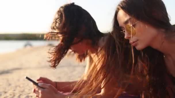 กลุ่มหญิงสาวมิลเลนเนียลใช้สมาร์ทโฟนวางอยู่ด้วยกันบนผ้าขนหนูชายหาดใกล้ทะเลในฤดูร้อนพระอาทิตย์ตกดิน ผู้หญิงสาวที่ติดโทรศัพท์มือถือสมาร์ท การสื่อสารผ่านอินเทอร์เน็ตตลอดเวลา . — วีดีโอสต็อก