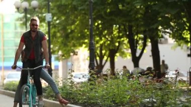 Parkta bisiklete binen genç bir çift. Romantik erkek arkadaş, mutlu kız arkadaşıyla şehir caddesinde kargo bölümünde bisiklet sürüyor. Kadınlar bisikletle gezerken erkeklere sarılır ve eğlenir. Yavaş çekim.