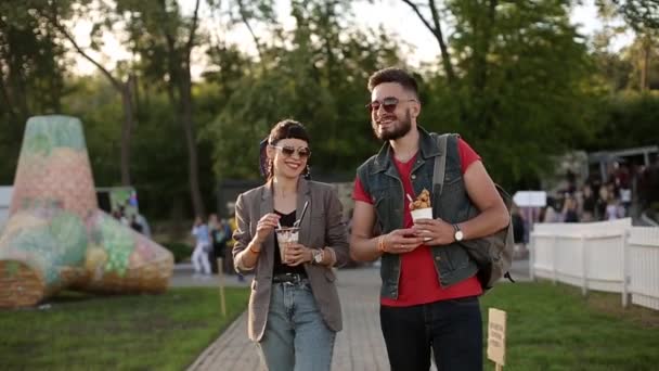 Stylowa hipsterska para chodząca po lodach i gofrach śmiejących się w parku miejskim. Romantyczny młody mężczyzna i kobieta bawiący się na świeżym powietrzu w czasie wolnym. Chłopak i dziewczyna w zwolnionym tempie. — Wideo stockowe