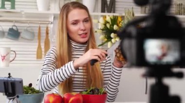 Yemek blogcusu mutfak stüdyosunda taze meyve salatası pişiriyor video kanalı için özel ders kameraları önünde çekim yapıyor. Kadın nüfuzlu kişi elmayı, ananası tutuyor ve sağlıklı beslenmekten bahsediyor. Fructorianism