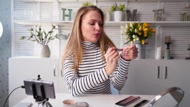 Vlogger samice nanáší rtěnku na kůži. Krása blogger žena natáčení denní make-up rutinní návod na kameru na stativ. Influencer blondýny dívka live streaming kosmetika recenzi produktu v domácím studiu — Stock video