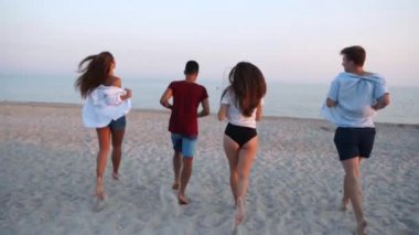 Bir grup mutlu arkadaş deniz suyuna doğru koşar ve plaja tişört atarlar. Neşeli insanlar okyanus kıyısında eğleniyorlar. Genç erkekler ve kadınlar yüzmeye ve banyo yapmaya giderler. 120 fps yavaş çekim.