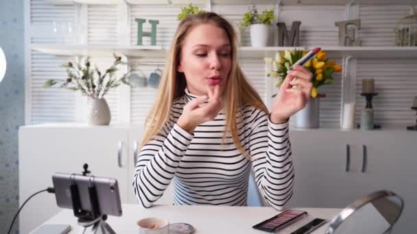 Vlogger kvinnliga applicerar läppstift på läpparna. Skönhetsbloggare kvinna filmar daglig makeup rutin handledning vid kameran på stativ. Influencer blond dam live streaming kosmetika produktrecension i hemstudio — Stockvideo