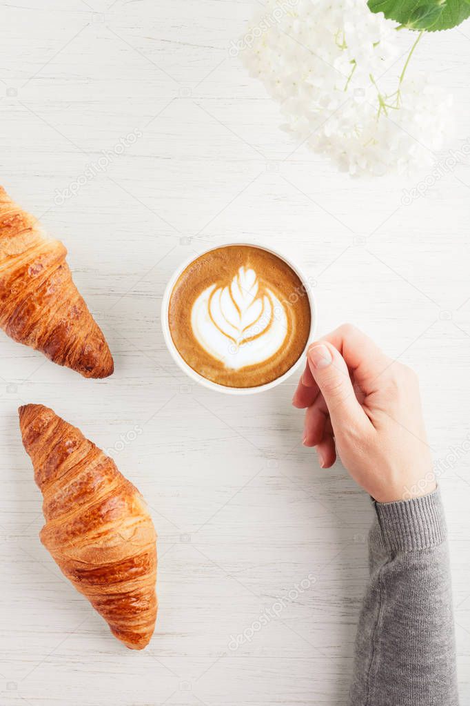 A cup of cappuccino with latte art and two croissants on white wooden table. Traditional french breakfast. Womans hand holding a cup of coffee. Top view.