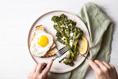 Kızarmış brokoli, limon, tabakta tost ve peçeteyle süslenmiş yumurta. Sağlıklı yiyeceklerin üst görüntüsü. Çatal bıçakla kadın eli..