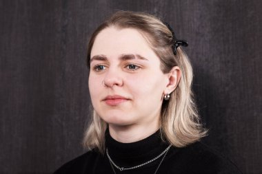 Kadın siyah bir balıkçı yaka kazak giyiyor ve saçında birkaç saç tokası var. Sol kulağında küçük bir küpe var. Arka plan koyu gri bir duvar..