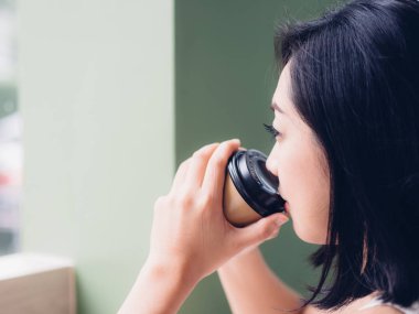 Asyalı kadın windows tarafından bir fincan sıcak kahve ve sit içiyor.