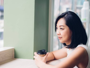 Asyalı kadın windows tarafından bir fincan sıcak kahve ve sit içiyor.