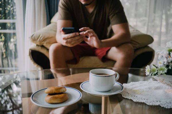 Asian man use smartphone and relax with coffee and cookies.