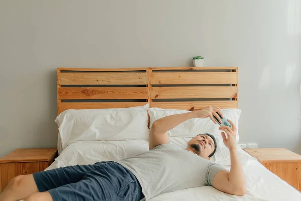 Homem está jogando jogo móvel com seu smartphone na cama . — Fotografia de Stock